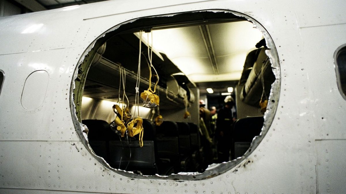 A white aircraft fuselage panel with a large, jagged-edged oval hole, through which yellow oxygen masks hang over rows of dark seats while figures in hard hats stand in the lit cabin beyond.