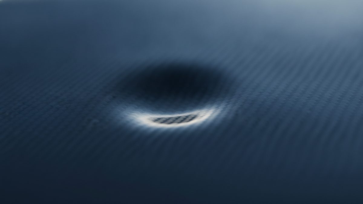 A close-up of a dark blue surface with a fine woven texture and a small, shallow circular dent near the center, its lower rim catching a crescent of white light that shows the weave inside the depression, with the rest of the surface softly out of focus.