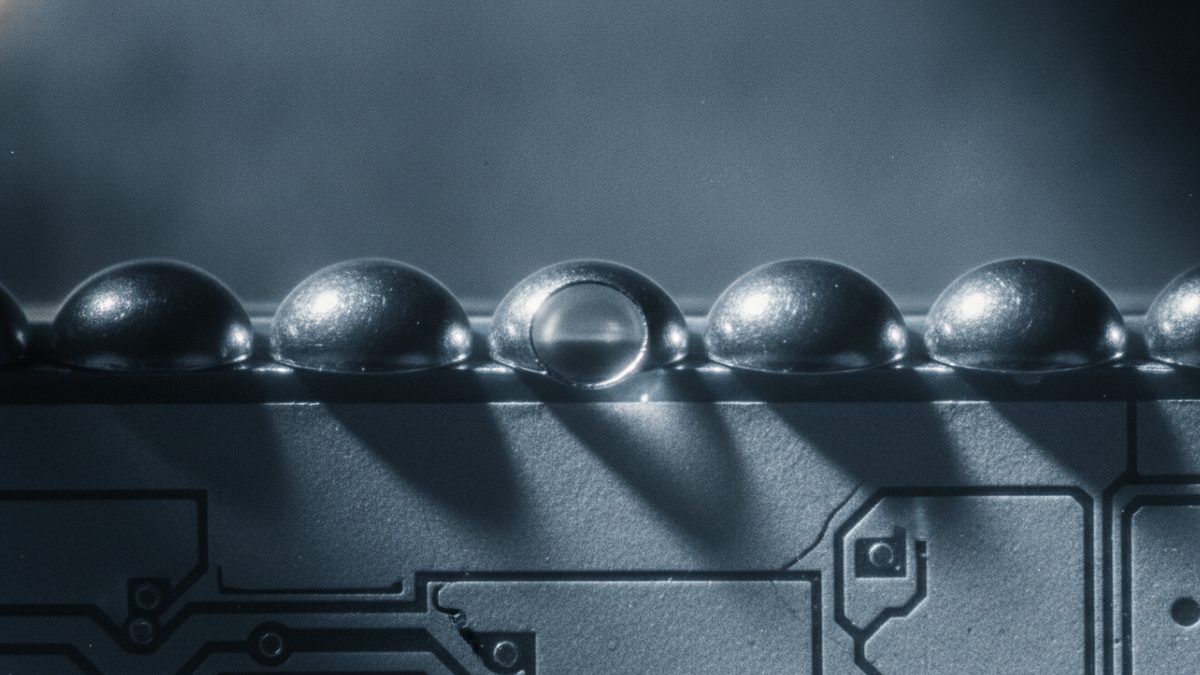 A close-up of a row of shiny dome-shaped metal balls along the edge of a gray printed circuit board, the middle one with a clear glassy disc set into its face, casting long diagonal shadows across the board’s traces, pads and a hairline crack against a blurred blue-gray background.