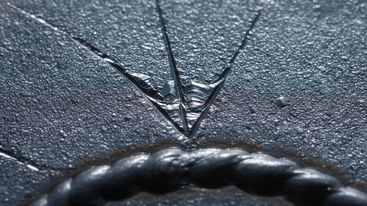 A macro close-up of a dark gray, finely granular steel surface in which a V-shaped crack with bright, faceted edges converges to a point just above a rippled, rope-like weld bead curving along the bottom of the frame, with thinner straight cracks running diagonally across the left side.