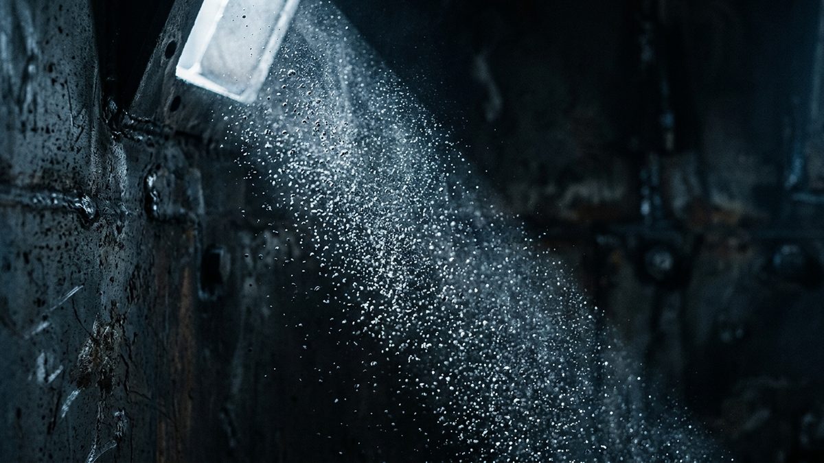 A shaft of light from a small rectangular opening at the upper left picks out a stream of fine particles drifting diagonally through a dark interior lined with weathered, rust-streaked metal walls and weld seams.