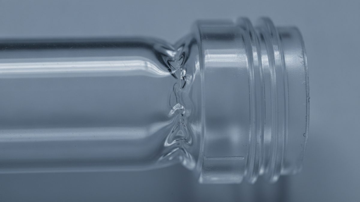 A macro photograph of a clear, empty plastic tube lying on its side against a pale blue-gray background, with a threaded, flanged neck at the right and a rippled, pinched shoulder where the neck meets the narrower body.