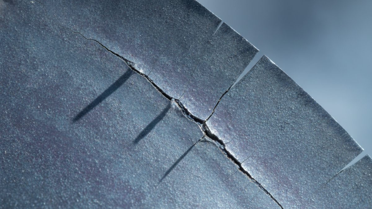 A macro close-up of a speckled blue-gray metal surface with a curved, notched edge at the upper right, split by a jagged dark crack running diagonally with fine branch cracks and thin shadow lines falling across it, against a soft blurred gray-blue background.