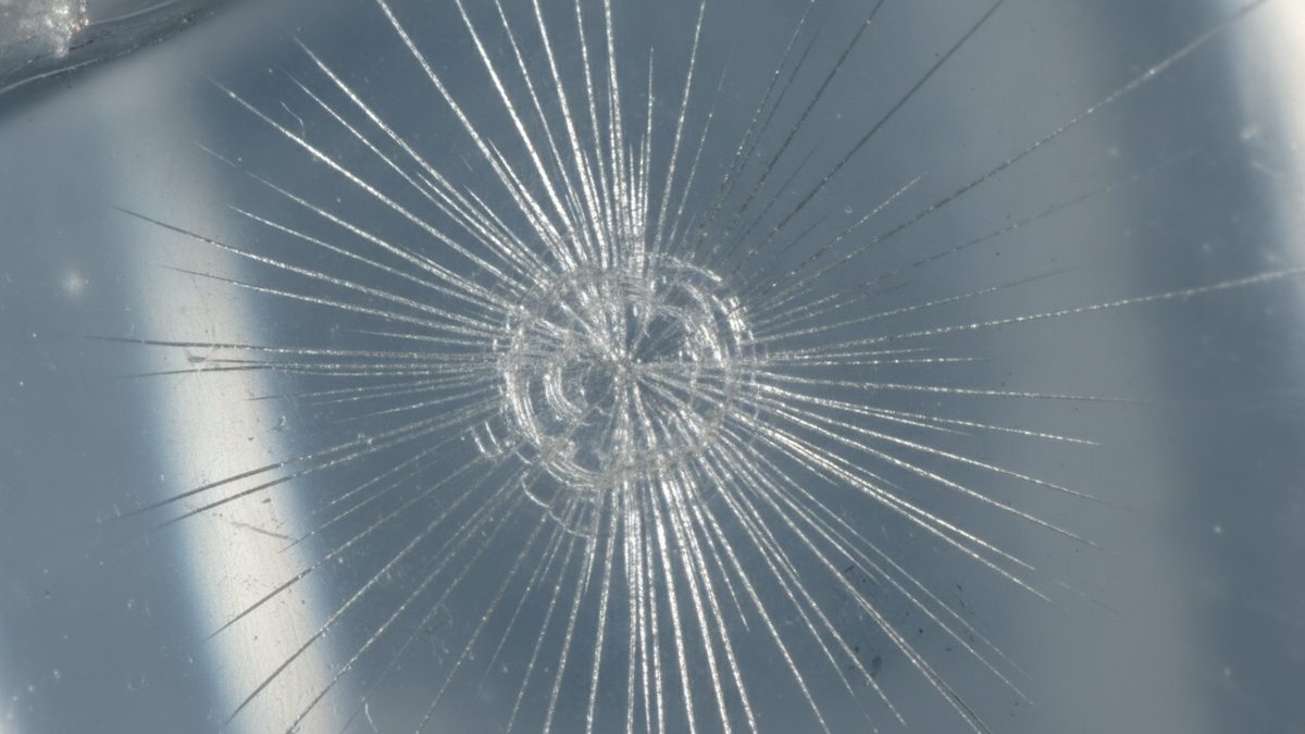 A macro photograph of a clear plastic surface with a starburst fracture: a small circular zone of concentric cracks at the center, with dozens of fine, straight cracks radiating outward in every direction against a blurred blue-gray background crossed by pale vertical bands of light.