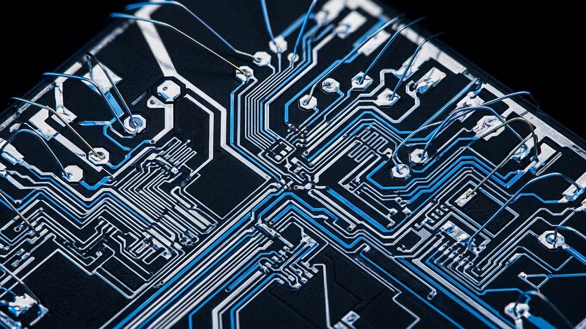 A close-up, angled view of a black circuit surface patterned with silver and blue metal traces, with thin arched wires bonded to round pads along its edges, set against a black background.