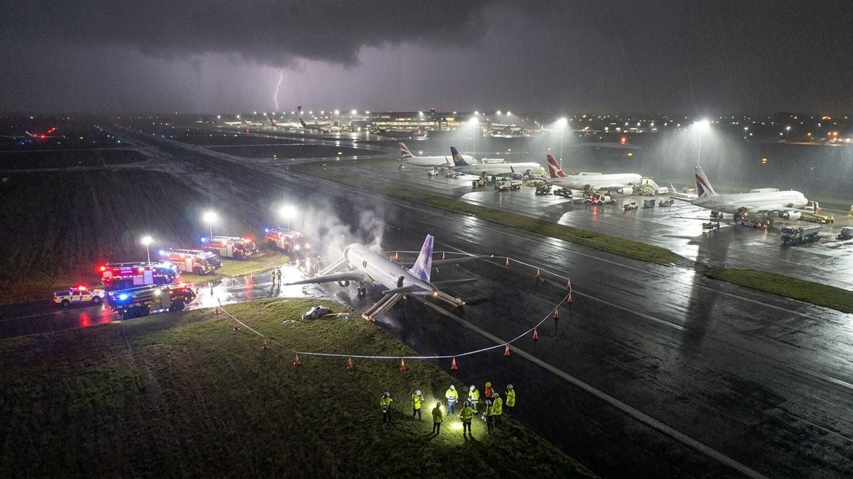 An aerial night view of a rain-soaked airfield under a stormy sky split by lightning, where a white jet with inflated evacuation slides sits inside a ring of orange cones and tape, fire engines with flashing lights and drifting smoke stand to its left, workers in high-visibility vests gather on the grass in the foreground, and parked airliners fill a floodlit apron in the distance.