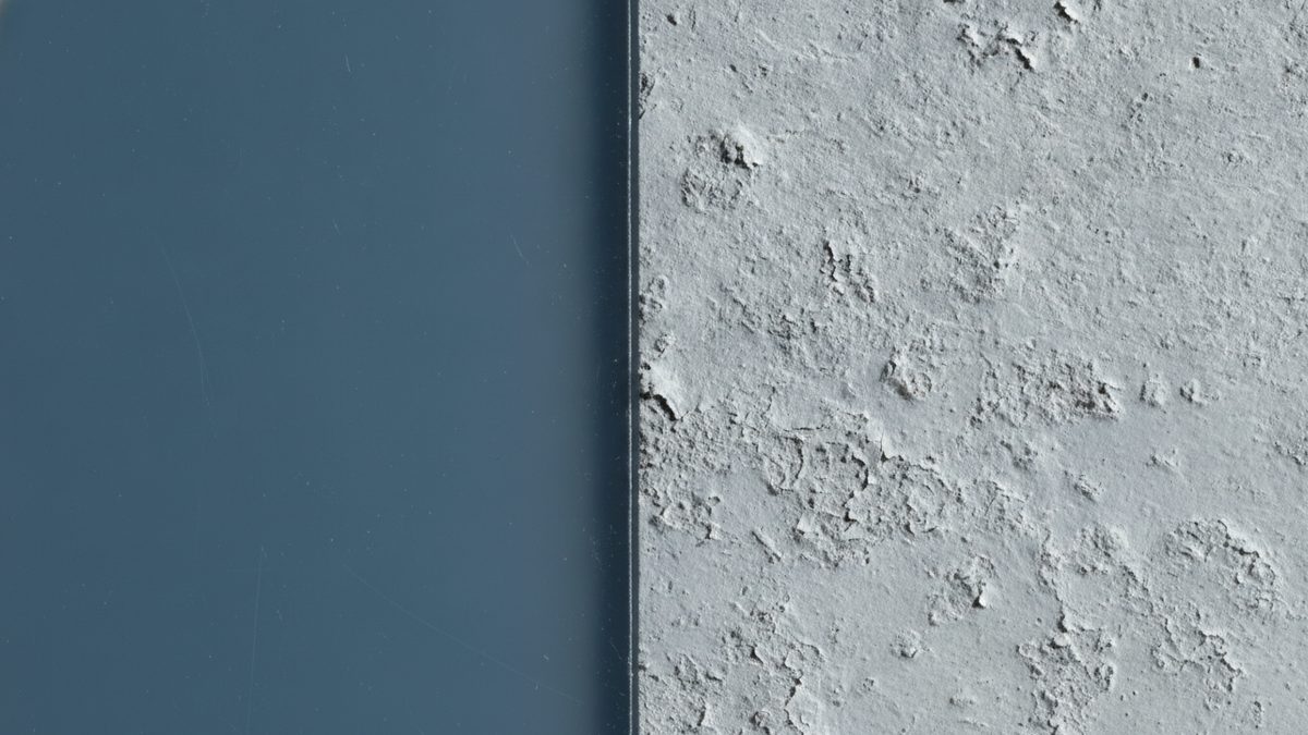 A close-up split view in which a smooth, glossy blue-gray plastic surface with faint fine scratches on the left meets, along a sharp vertical edge, a rough pale gray surface on the right that is crusted, pitted and flaking.