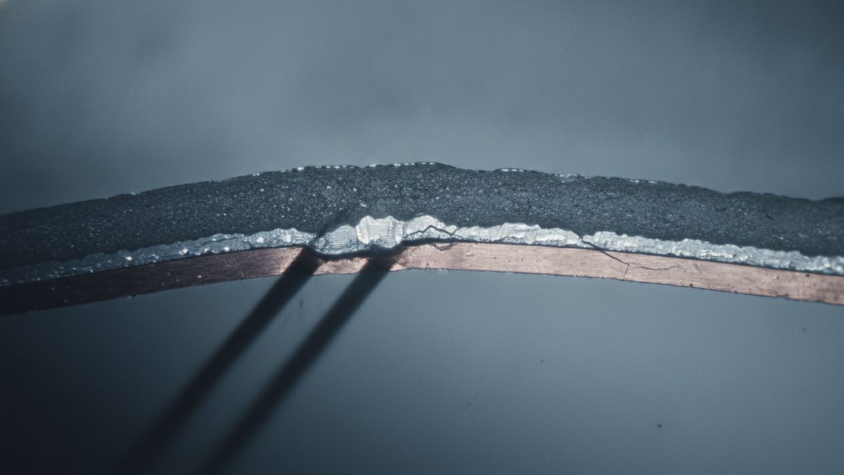 A close-up of a curved, layered strip — a dark gray granular top layer over a wavy silvery layer and a copper band — with fine cracks through the silvery layer near a slight kink at its center, set against a blurred blue-gray background crossed by two dark diagonal shadows.