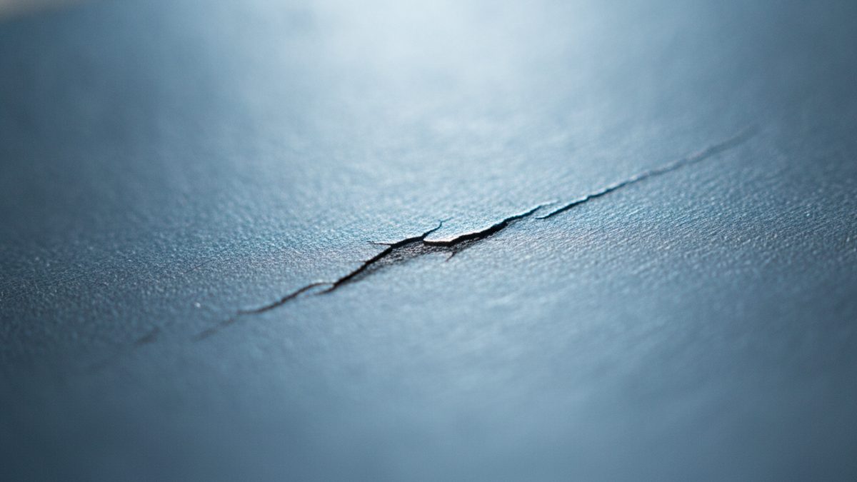An extreme close-up of a finely textured blue-gray surface with a single thin, jagged dark crack running diagonally across the center, the crack in sharp focus while the foreground and background fall into soft blur under pale light from the top of the frame.