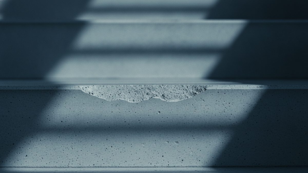 A close-up of gray concrete stairs in cool blue light, with a chunk broken away from the leading edge of the nearest step to expose rough, pitted concrete, while diagonal bands of light and shadow fall across the steps and the upper steps blur into the background.