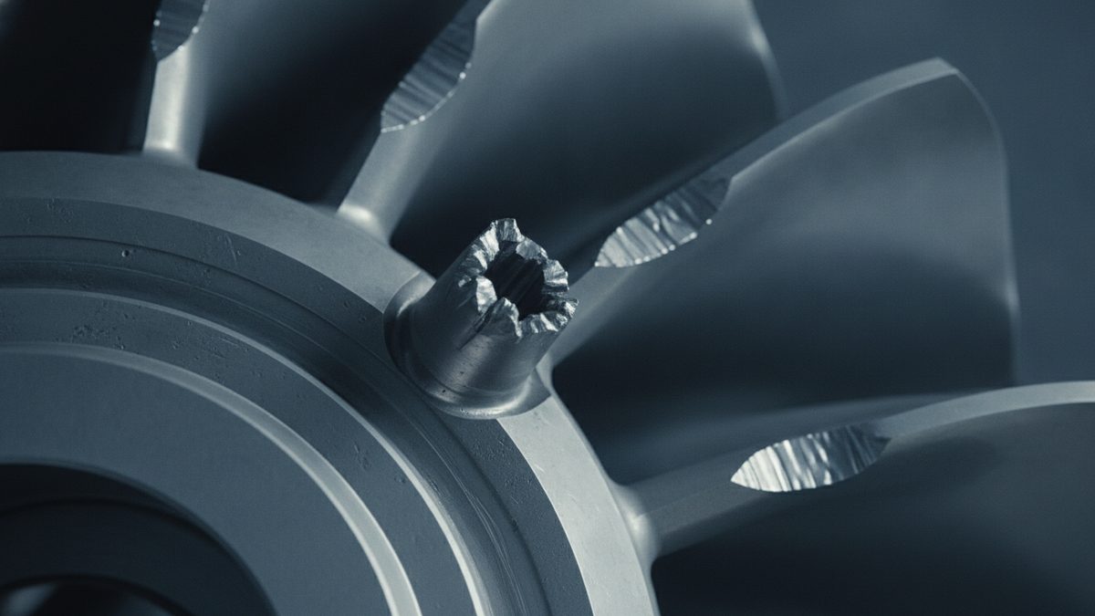 A close-up, blue-gray view of a bladed metal rotor disc, with concentric rings on its hub at left and several radiating blades showing jagged, silvery fracture surfaces, including a short hollow stub with a torn, crumpled rim projecting from the hub.